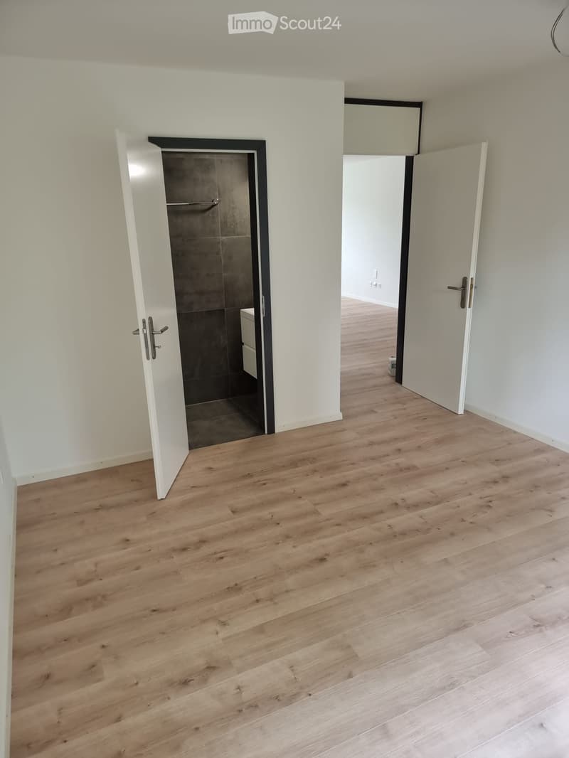Empty room with light wooden flooring and white walls. Open doors lead to a bathroom and another room.