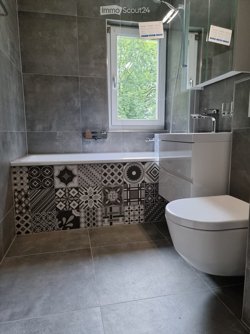Modern bathroom with grey tiled walls, a bathtub with decorative black and grey tiles, a toilet, a sink with a modern faucet, and a glass-enclosed shower area.