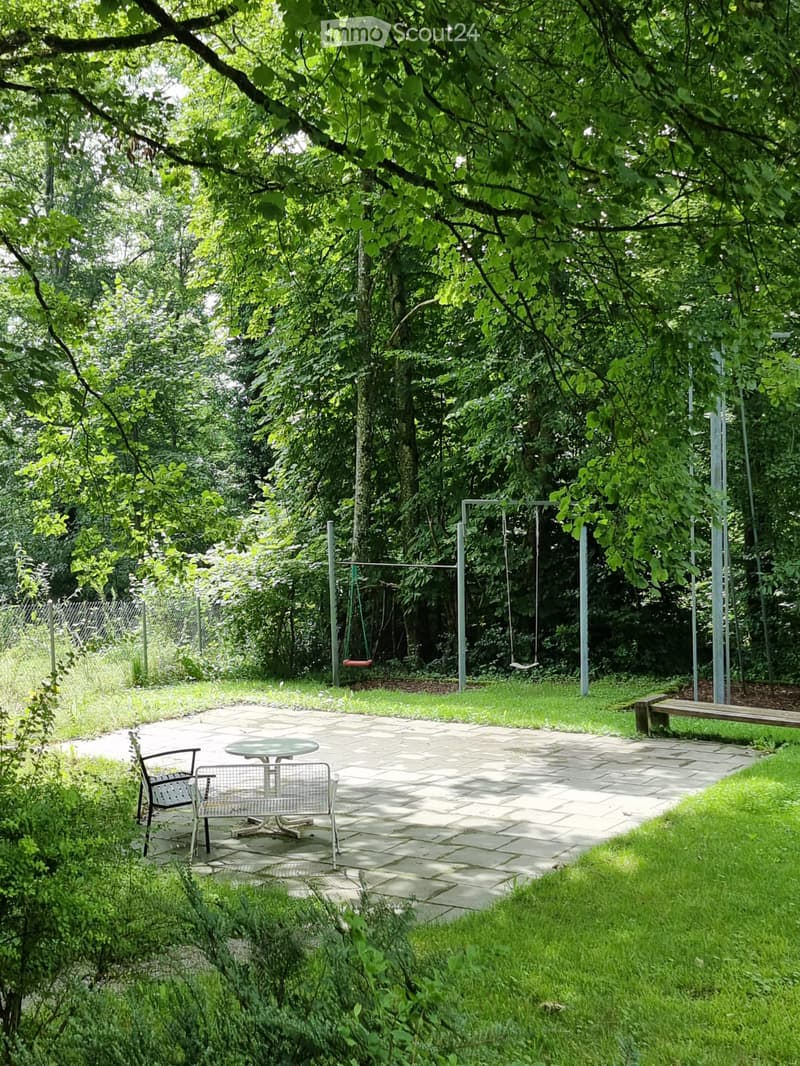 A serene garden area with a stone-paved patio, a round table with two chairs, a wooden bench, and a swing set surrounded by lush greenery.