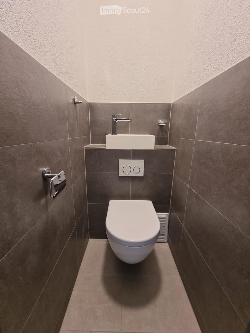 Modern bathroom with a toilet, wall-mounted sink, and sleek faucet. Grey tiled walls and floor.