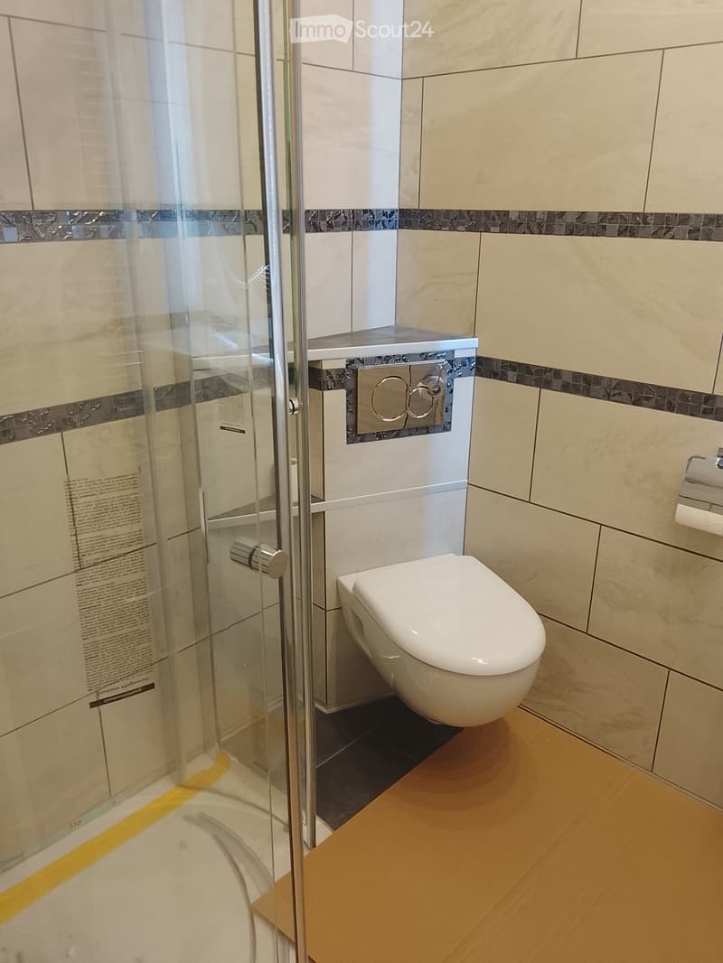 Modern bathroom with a white toilet, glass-enclosed shower stall, beige tiles, and a dark grout border.
