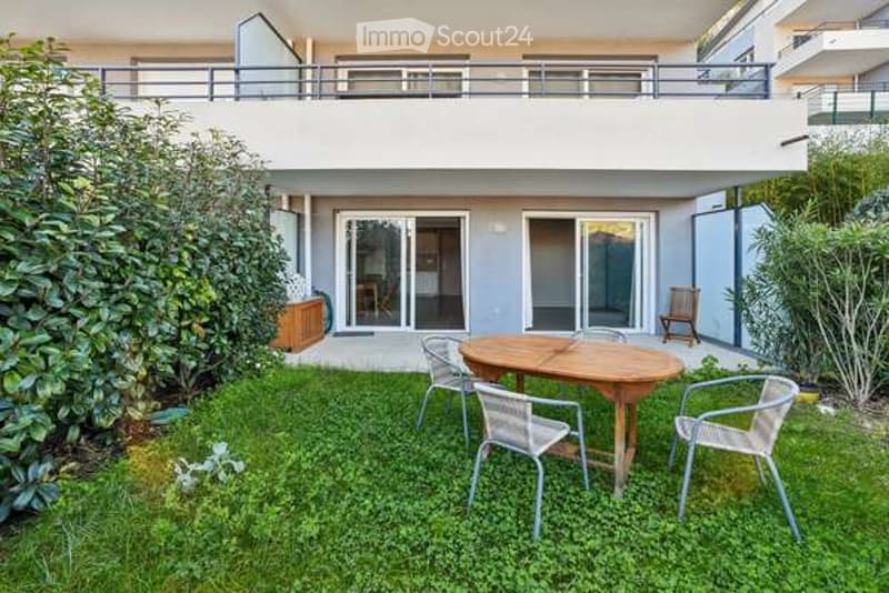 Well-maintained garden area with a wooden round table, four metal chairs, and a small wooden bench. The area is surrounded by green grass and shrubs, with a modern building featuring a balcony and large glass doors in the background.