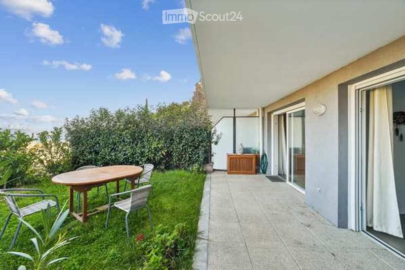 Balcony with a small grassy area, a wooden table, and four metal chairs. The balcony is connected to an indoor space with a small wooden cabinet and a view of the surrounding greenery.