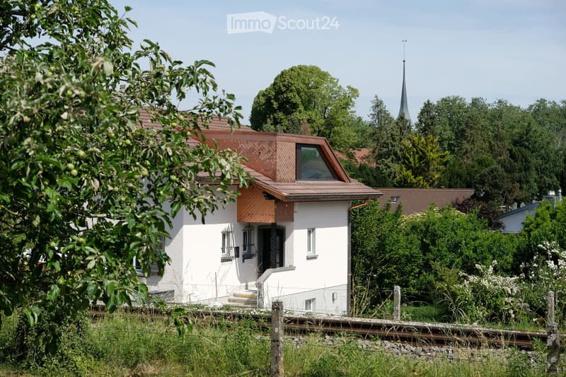 A modern single-story house with white walls, a red-tiled roof, and a small balcony. The house is surrounded by lush greenery and trees, with a visible church spire in the background.