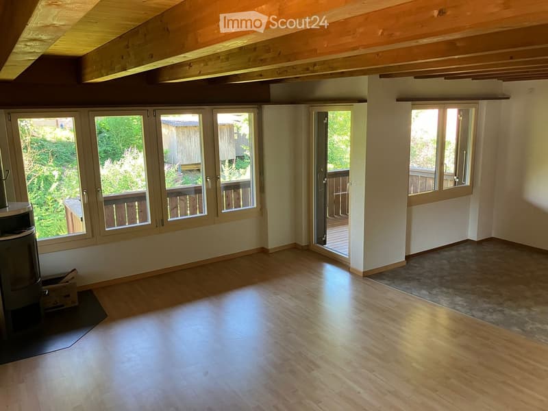 Bright, empty room with wooden flooring, large windows, and a sliding door leading to a balcony. Exposed wooden beams on the ceiling.