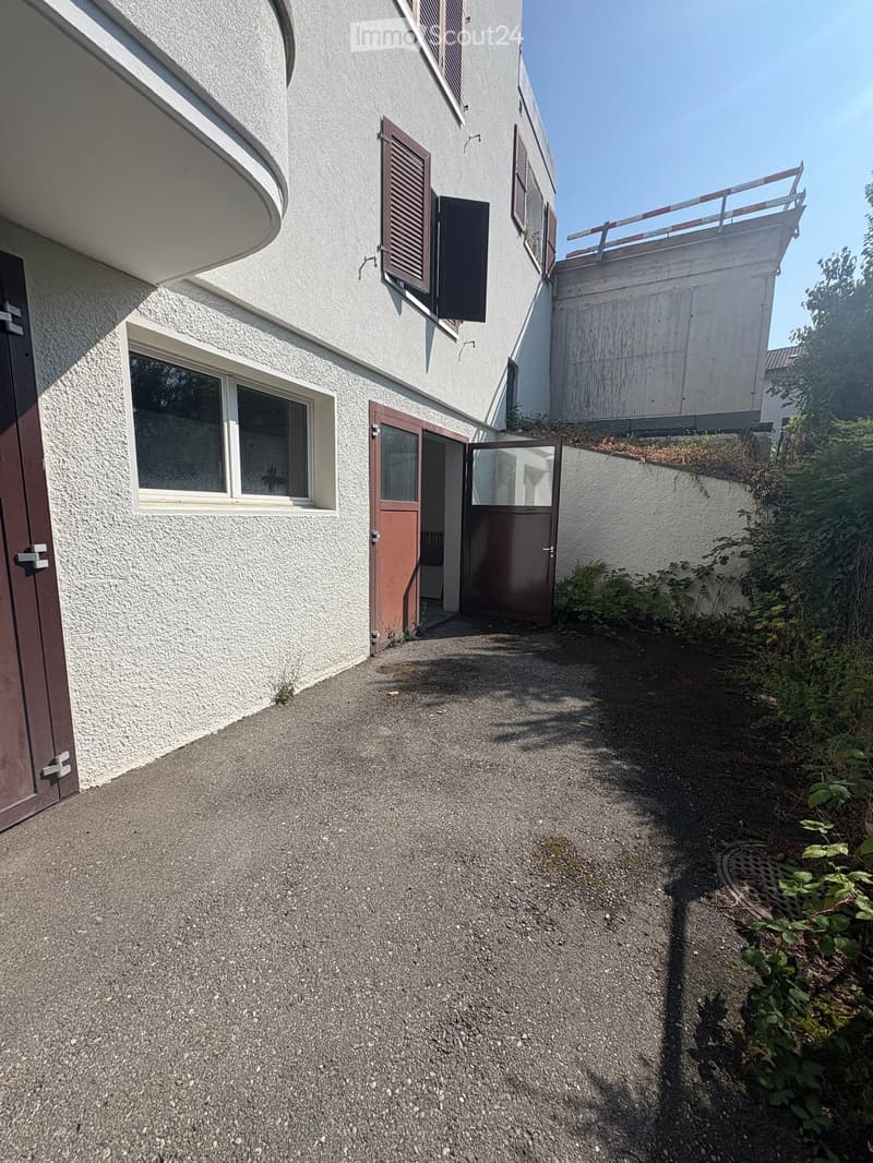 Narrow gravel pathway leading to a building entrance, with a small garden area on the right side and a raised section with a railing on the left.