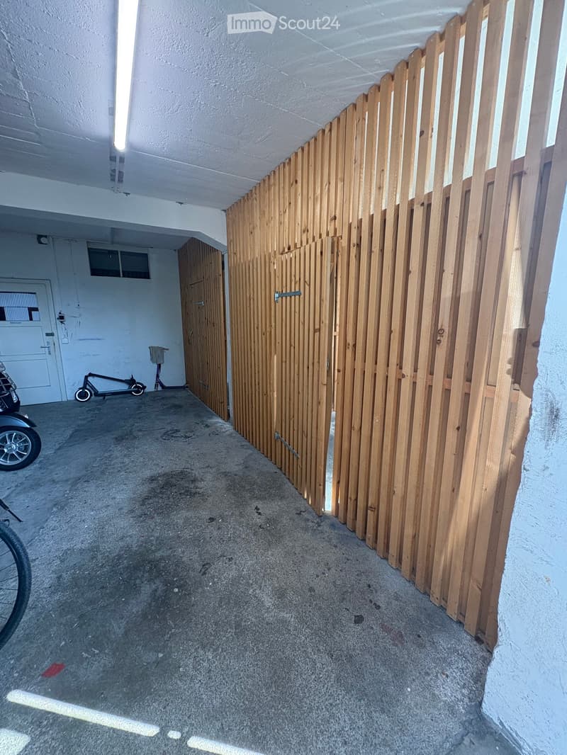 A narrow, enclosed corridor with a wooden slatted fence on one side, concrete floor, and a few parked vehicles including a scooter and a bicycle.