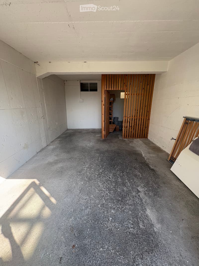 Empty concrete-floored corridor with white walls, a small window, and a wooden door leading to another room.