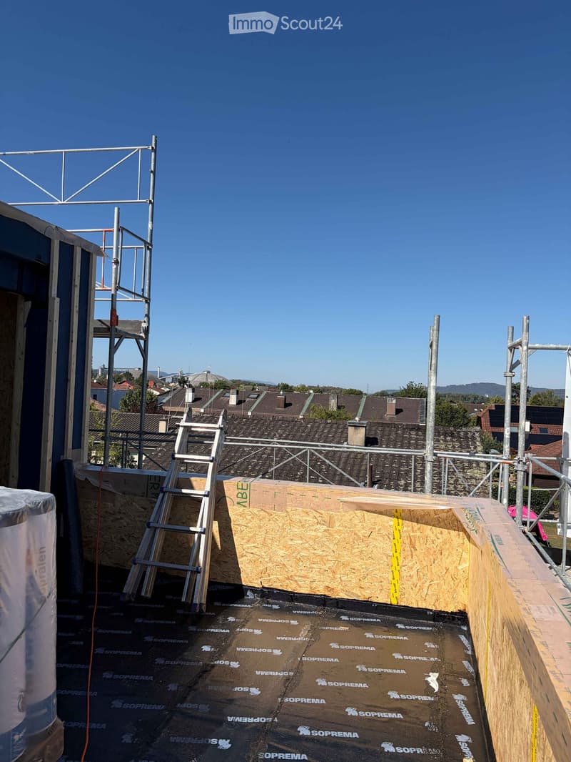 Roof under construction with wooden framing, insulation material, and scaffolding. The background shows a cityscape with rooftops and distant hills.
