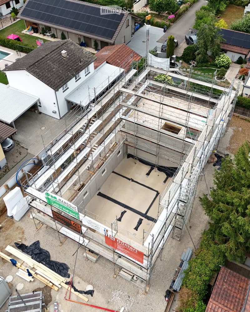 Aerial view of a residential area with multiple houses, some under construction with scaffolding and exposed concrete floors, surrounded by greenery and parked cars.