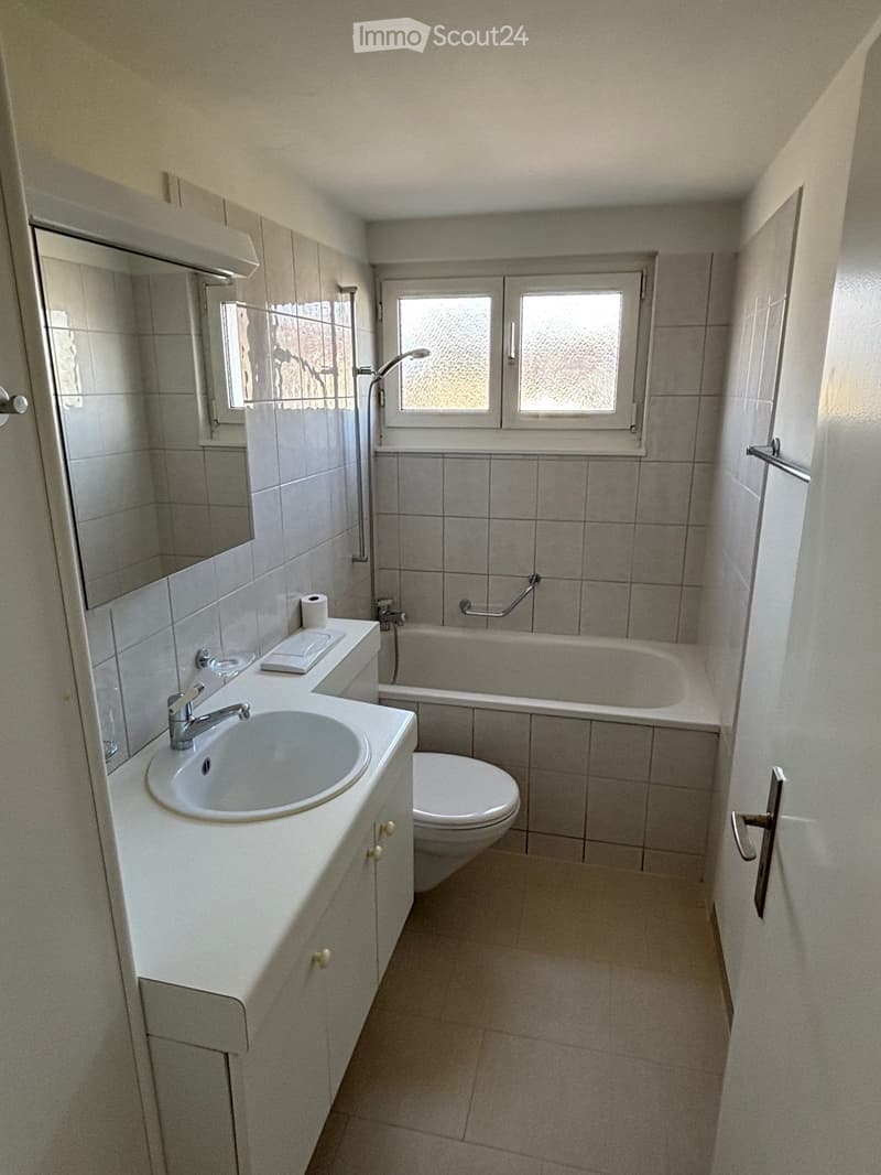 Modern bathroom with a white sink, mirror, toilet, and bathtub. The room features tiled walls and a window with frosted glass panes.