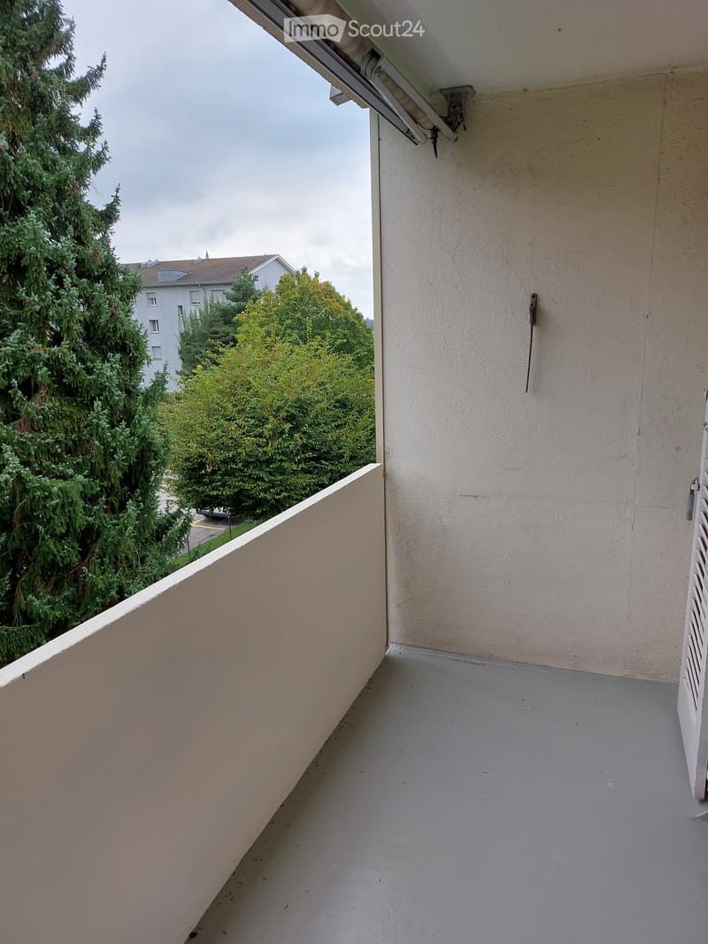 Balcony with a low railing, grey floor, and beige wall, overlooking a green area with trees and a building in the background.