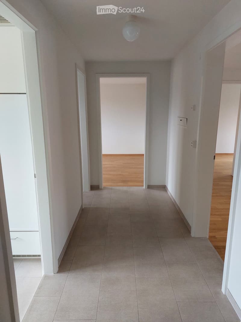 Empty hallway with tiled floor and wooden flooring visible in adjacent rooms, white walls, and a ceiling light fixture.