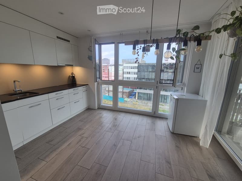 Modern kitchen with white cabinets, wooden flooring, and large windows offering a city view. Features a sink, faucet, and a refrigerator.