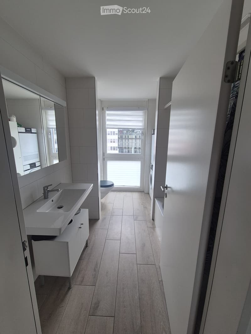 Modern bathroom with a white sink, faucet, mirror, and cabinet. The floor is light wood, and a toilet is visible in the background. A large window provides natural light.