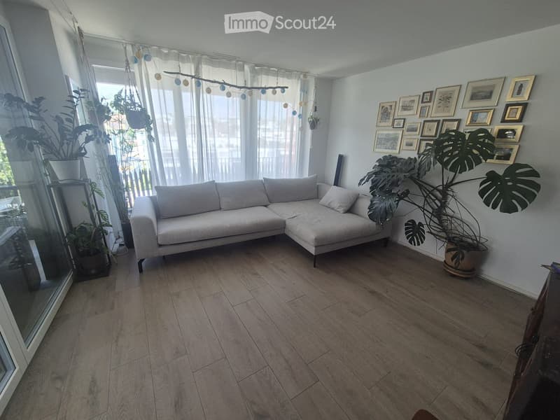 Modern living room with light-colored wooden flooring, a grey sectional sofa, large potted plants, and a wall decorated with framed pictures.