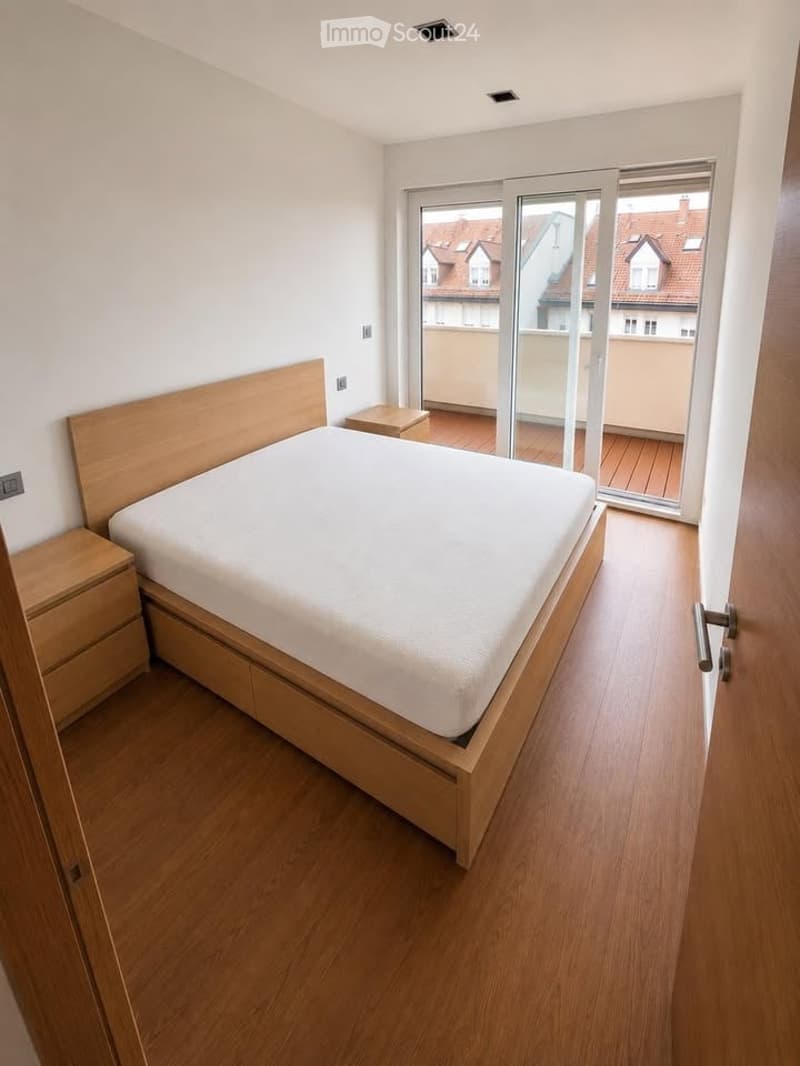 Modern bedroom with wooden flooring, a large bed with white bedding, a nightstand, and a sliding glass door leading to a balcony.