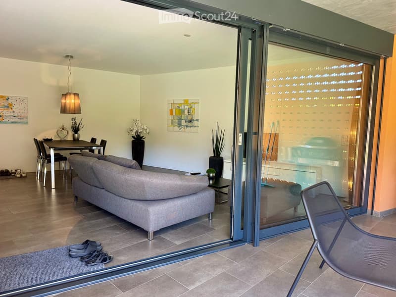 Modern living room with a grey sofa, coffee table, dining table, chairs, pendant lamp, artwork, and vases. Large sliding glass doors lead to a balcony area with a chair.
