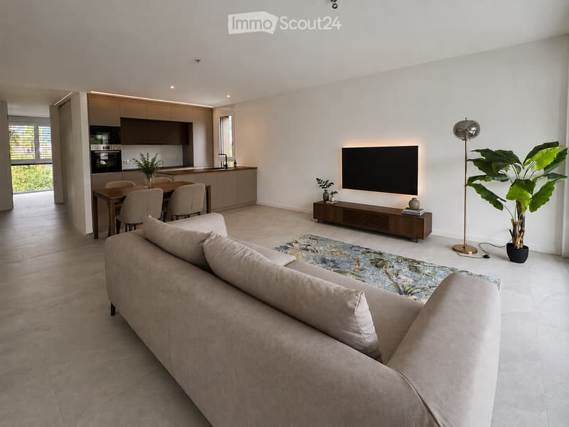 Modern living and dining area with a beige sofa, coffee table, dining table with chairs, flat-screen TV, floor lamp, and a large plant. The kitchen area features cabinets, an oven, and a faucet.