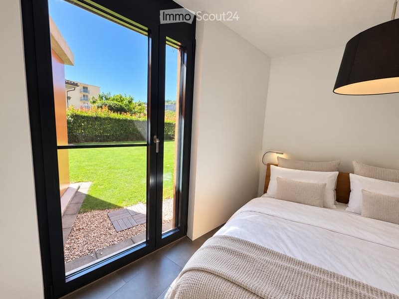 Modern bedroom with a large bed, white bedding, and a view of a well-maintained garden through large glass doors.
