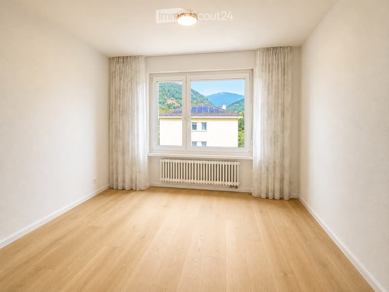 Empty room with light wooden flooring, white walls, large window with curtains, radiator, and a view of a house with solar panels on the roof.