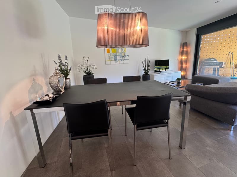 Modern dining and living area with a sleek black dining table, chairs, a grey sofa, a hanging lamp, and decorative vases.