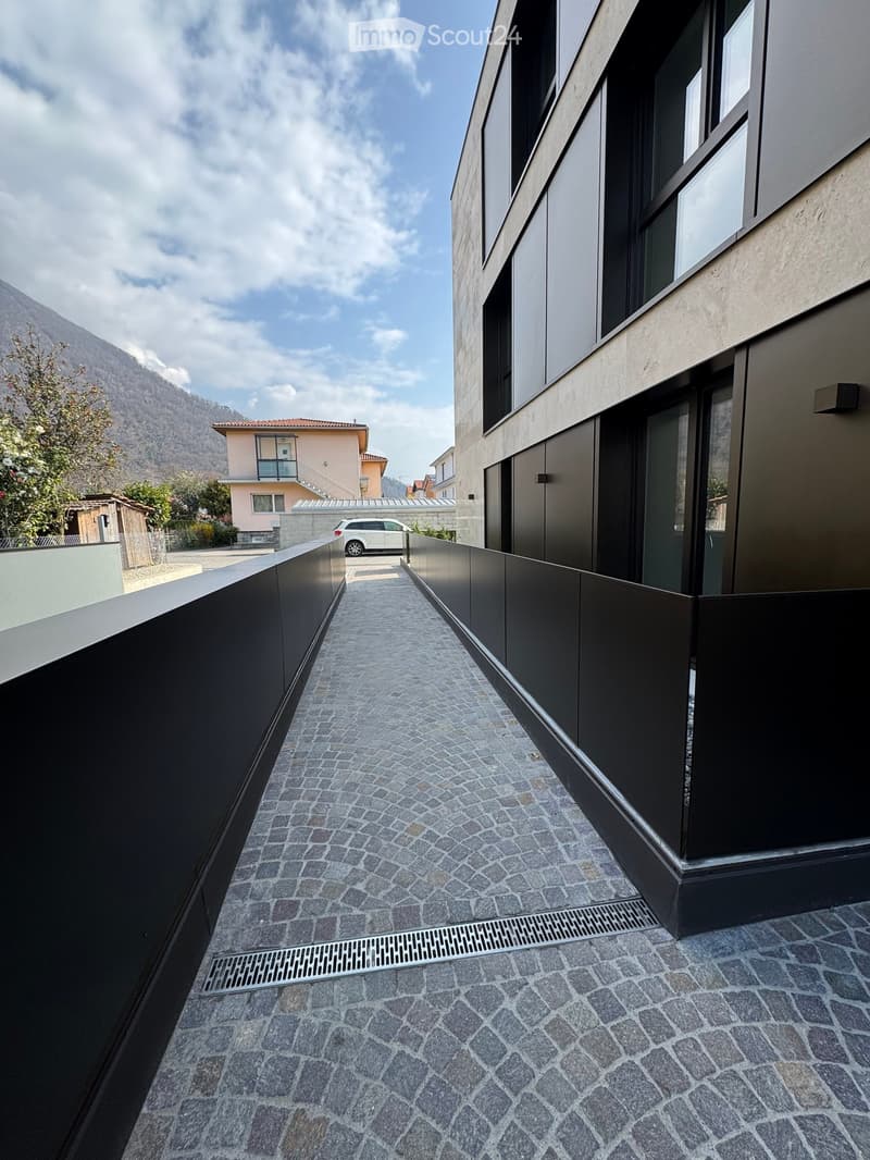 Corridor / Hallway. Narrow paved pathway between two modern buildings, with stone tiles and metal drainage grates.