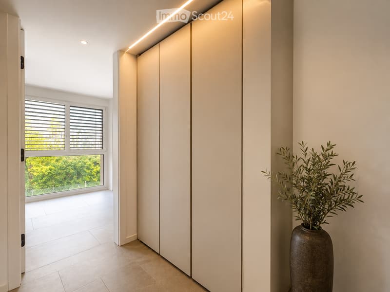 Modern hallway with white walls, recessed lighting, large window with blinds, and a tall decorative vase with greenery on the right side.