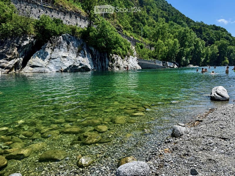 A serene river surrounded by lush green trees and rocky cliffs, with people enjoying the water and a stone bridge in the background.