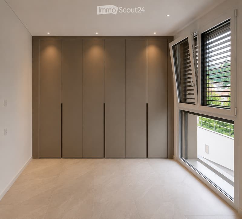 Empty room with beige tiled floor, large built-in wardrobe, and large windows with blinds, offering a view of greenery outside.