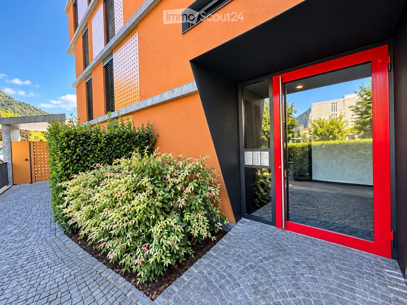 Modern building entrance with a red door, glass panels, and a small garden area with shrubs and a pathway.