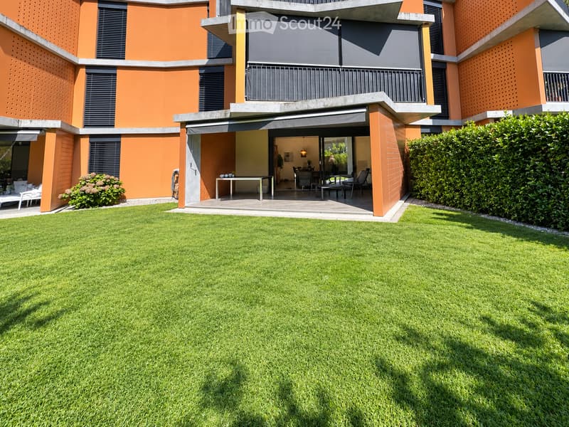 Modern house with orange and grey facade, large lawn, patio with outdoor furniture, and covered entrance area.