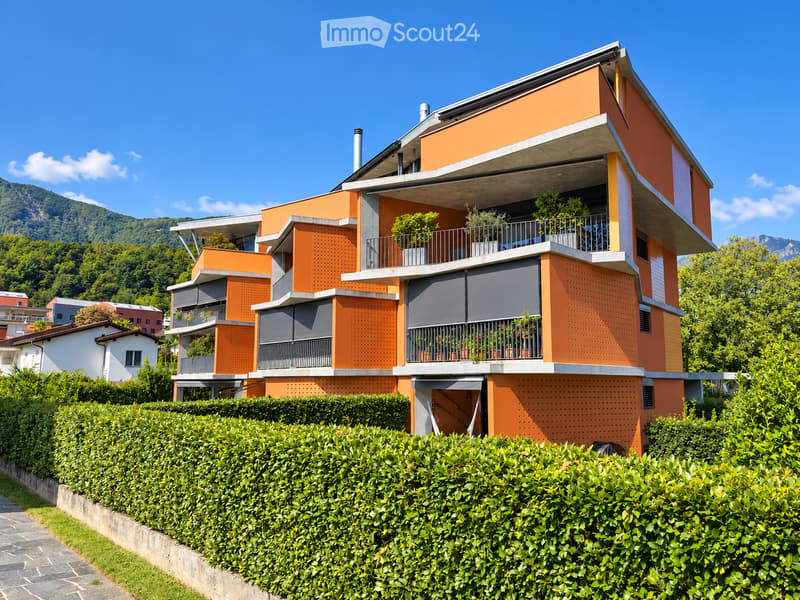 Modern two-story building with orange and gray facade, multiple balconies with potted plants, surrounded by greenery and a hedge.