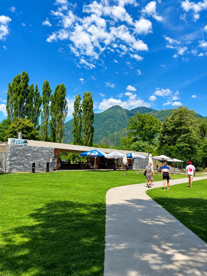 A well-maintained garden area with a paved pathway, green lawn, outdoor seating area with umbrellas, and a modern building labeled "RIVER" in the background. The scene is surrounded by tall trees and mountains under a clear blue sky.