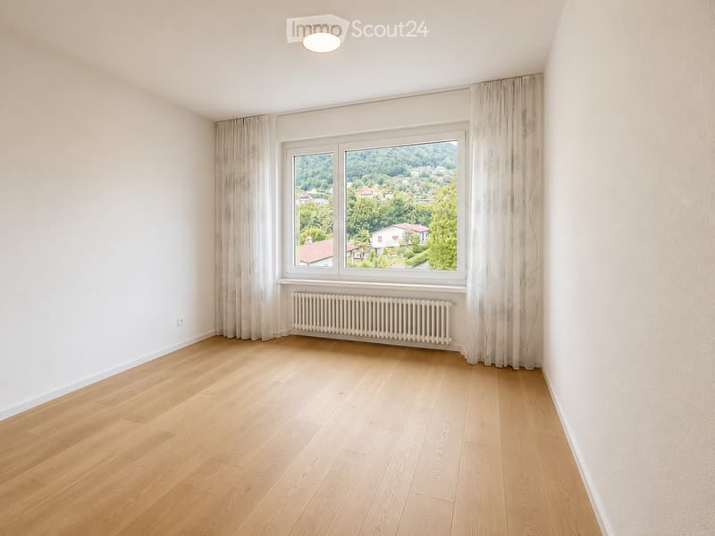 Empty room with light wooden flooring, white walls, large window with curtains, radiator, and a view of greenery and houses.