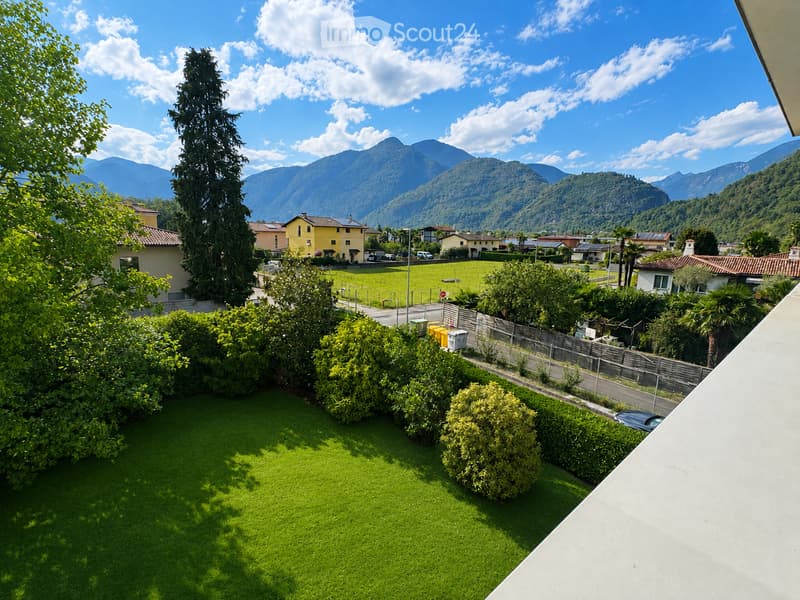 A well-maintained garden with green grass, trimmed bushes, and trees, overlooking a scenic valley with mountains and houses in the background.
