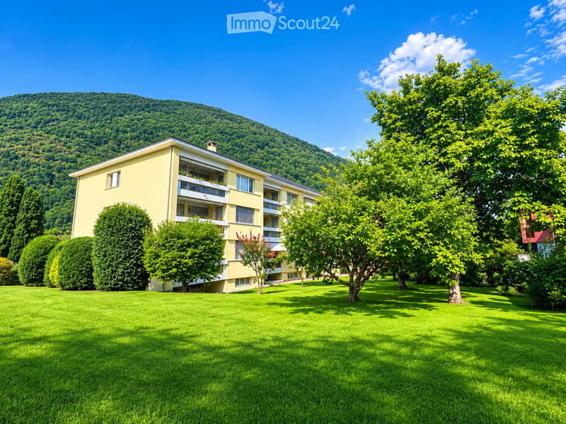 A yellow multi-story building with balconies, surrounded by a well-maintained lawn and trees, with a forested hill in the background.