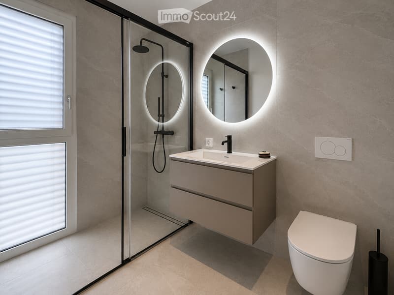 Modern bathroom with a sleek sink, round illuminated mirror, toilet, and a glass-enclosed shower area.