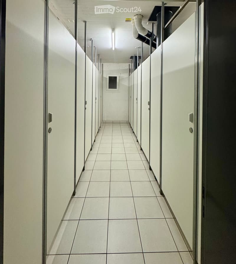 Clean, modern hallway with tiled floor and white walls, featuring multiple stall doors and a small window at the far end.