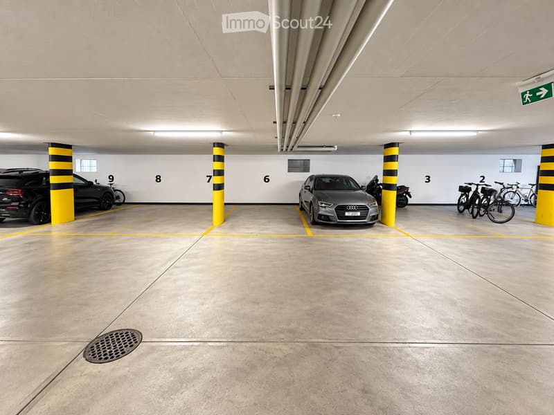 Underground parking area with marked parking spaces, several cars, bicycles, and a motorcycle. The area is well-lit with a clean, modern design and numbered parking spots.