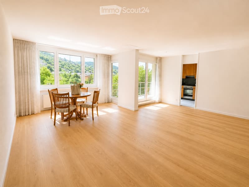Bright, spacious room with wooden flooring, large windows with curtains, a dining table with chairs, and a handbag with flowers on it. A kitchen area is visible in the background.