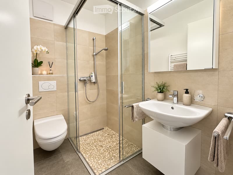 Modern bathroom with beige tiles, a glass-enclosed shower, a toilet, a sink with a mirror, and decorative items including a flower vase and a candle.