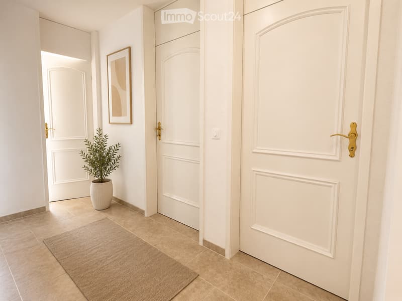 A clean, modern hallway with white walls, tiled flooring, and several closed white doors with gold handles. A small potted plant and a framed picture are placed on the left side.