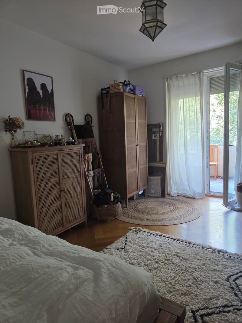 Cozy bedroom with a bed, wooden wardrobe, cabinet, mirror, picture frame, vase with flowers, and a large window with curtains leading to a balcony.