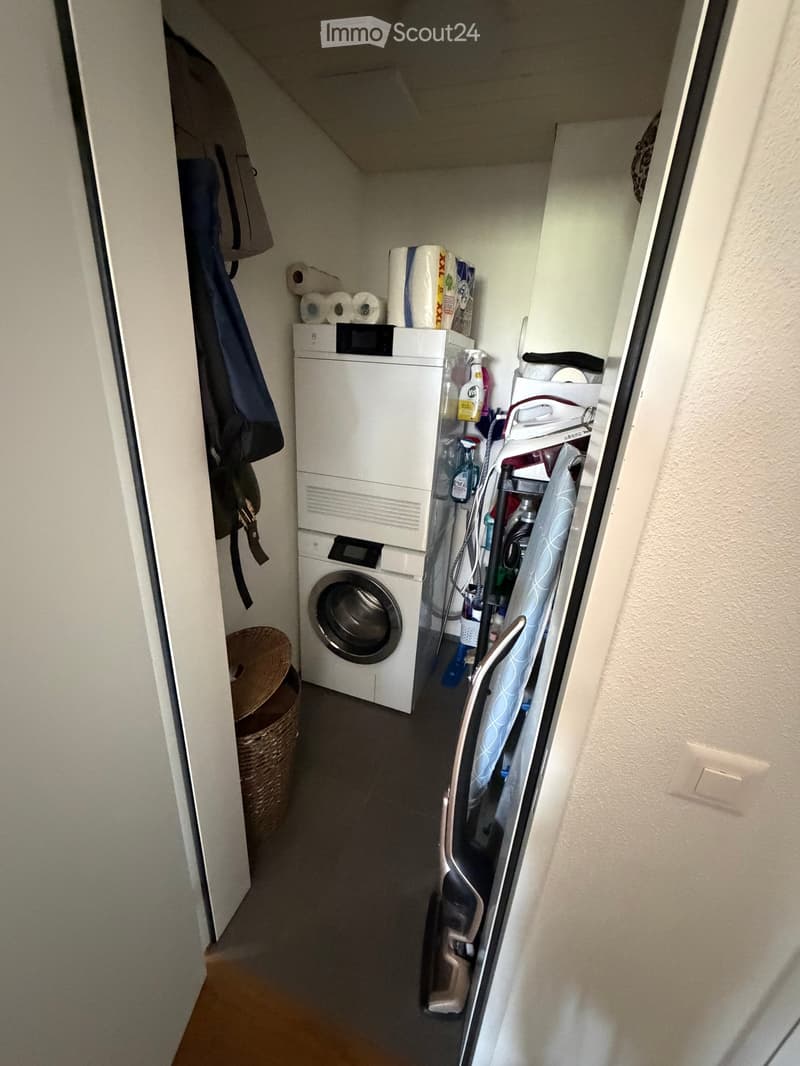 Laundry area with a stacked washer and dryer, toilet paper, cleaning supplies, and a wicker basket.