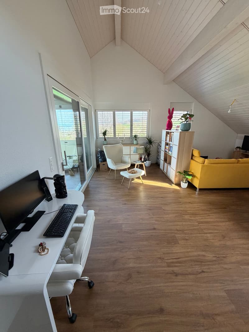 Bright living room with wooden flooring, large windows, a yellow sofa, a white armchair, a small table, a desk with a computer, and decorative items including a rabbit sculpture and plants.