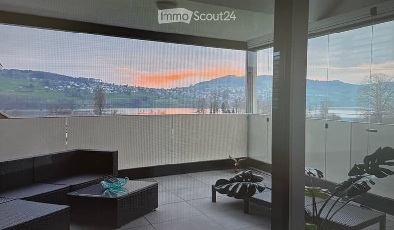 Balcony with large windows offering a scenic view, featuring a dark wicker sofa, a small table, a lounge chair, and a leafy plant.