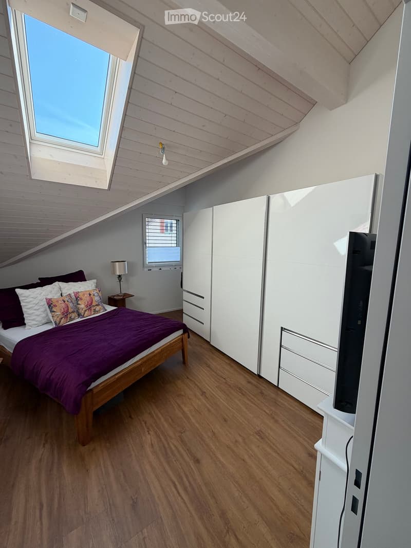 Cozy attic bedroom with wooden floors, slanted wooden ceiling, large window, bed with purple bedding, nightstand with lamp, white wardrobe, and TV on stand.