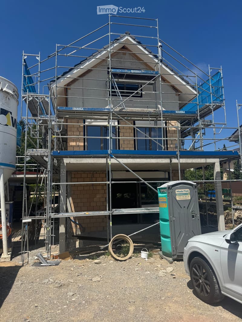 A two-story house under construction, covered with scaffolding, with visible brickwork and windows, surrounded by construction materials and a portable toilet.