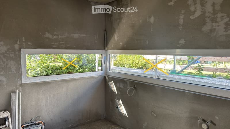 Empty room with unfinished concrete walls and two windows showing greenery outside. No furniture or equipment present.